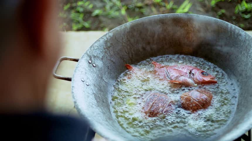 A whole reddishorange fish and two fillets are frying in a large metal pot of bubbling oil on a stovetop with a persons blurred shoulder seen in the foreground and a grassy outdoor area in the background