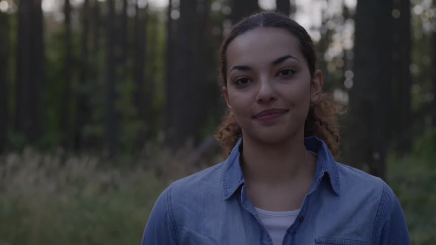 Bi racial pretty young girl is smiling as she looks affectiently at the camera she is pretty with well shaped eyebrows and is wearing a jean type button up shirt she is standing in the woods which appears to be around dusk