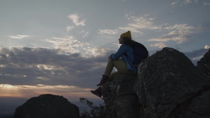 Ayoung woman carrying a backpack sitting on the edge of a rocky cliff as she looks out towards the sky