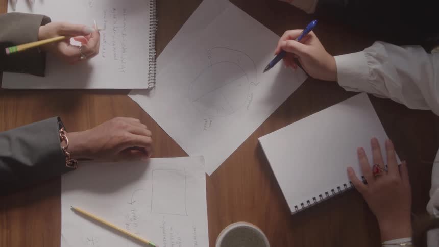 A topdown view shows several peoples hands some wearing rings and bracelets working together with pencils and a pen to draw diagrams on sheets of paper on a wooden table