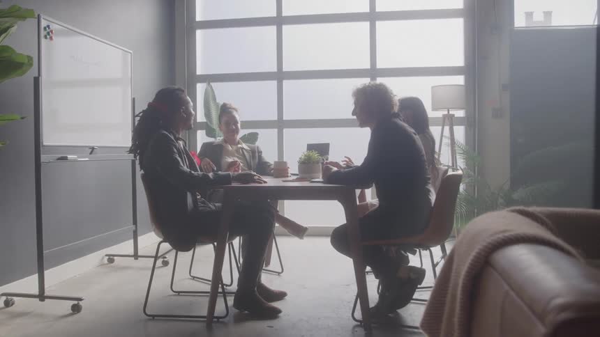 Four professionals are sitting around a meeting table in a modern office talking and gesturing with their figures silhouetted against a large brightly lit window behind them