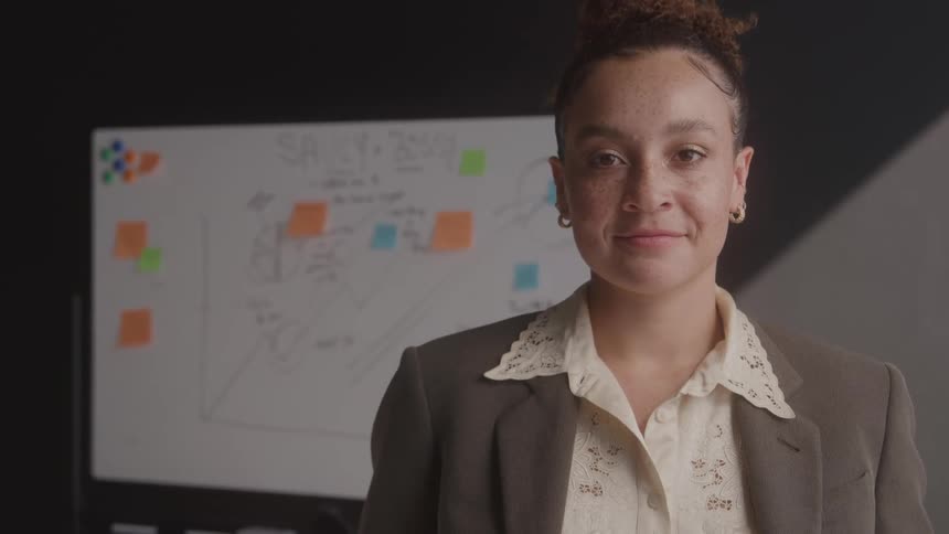 Woman wearing a brown blazer and light lace shirt standing in front of a whiteboard with notes and colorful sticky notes
