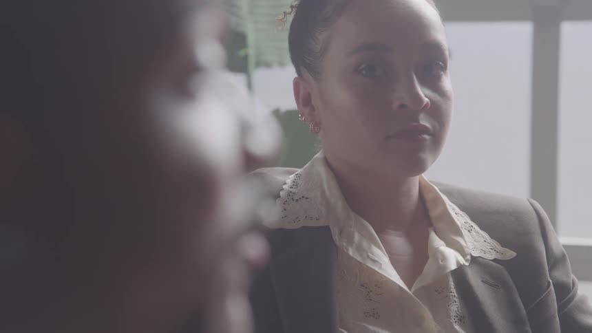 A woman with her dark hair pulled back wearing a blazer over a lace collared blouse is sitting and talking with a serious expression to another person in the room