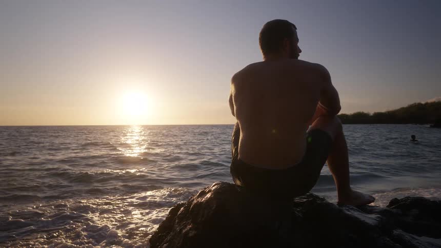 A man sitting on a small rock on a beach looks at the sun above the horizon then looks to his side