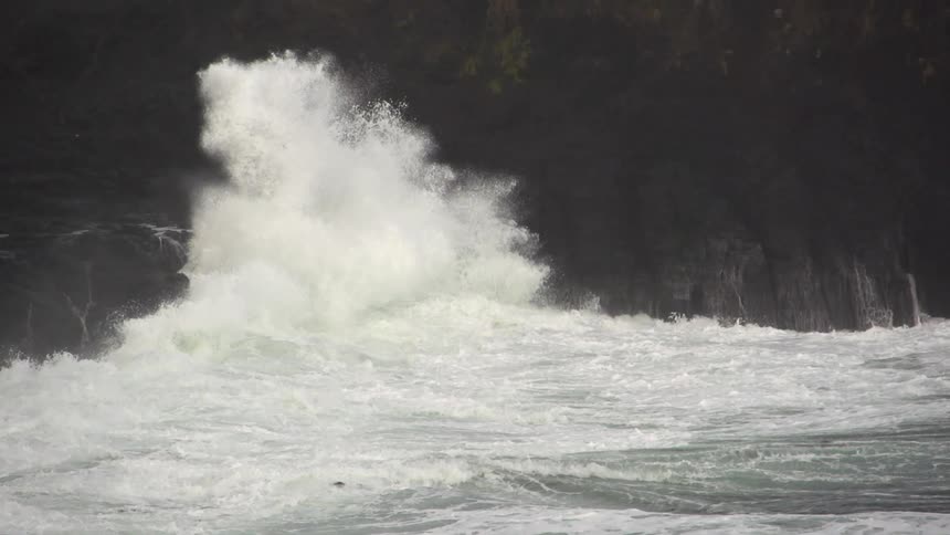 Large waves crashing onto a cliff, spraying upwards and water draining from the rocks back to the ocean