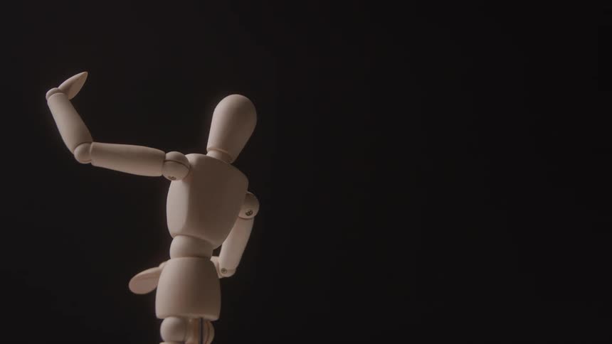 The video shows a wooden mannequin or artists model standing upright against a black background with its left arm raised the articulated figure is slowly turning its body showcasing its hinged joints