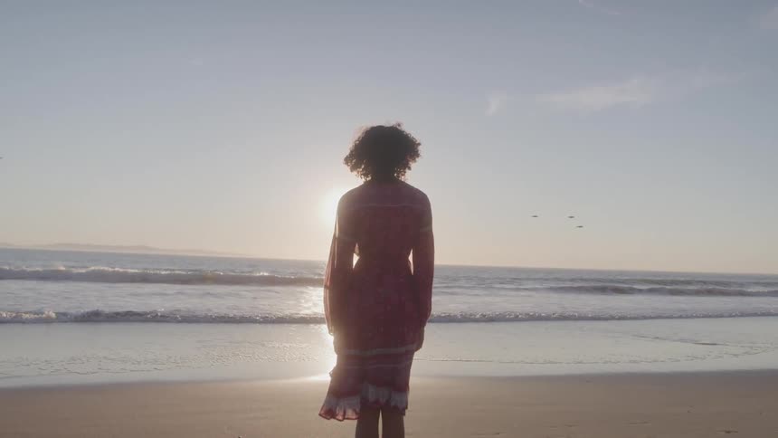 A young woman with curly hair stands on a beach facing the ocean the shot is a silhouette of her as the sun sets over the water creating a serene and contemplative mood