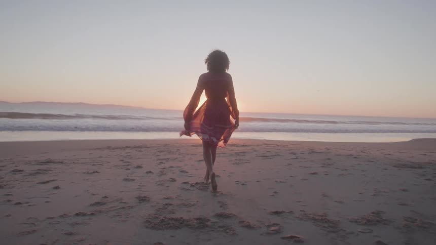 A woman in a flowing dress walks barefoot on the sandy beach towards the ocean