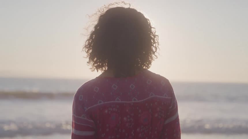 A young woman wearing a red dress stands on the beach watching the ocean waves hit the shore at midday