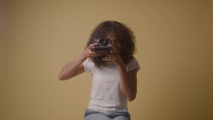 A woman with curly hair wearing a white tshirt stands in front of a plain background she holds an instant camera first aiming it at the viewer and then smiling after taking the picture