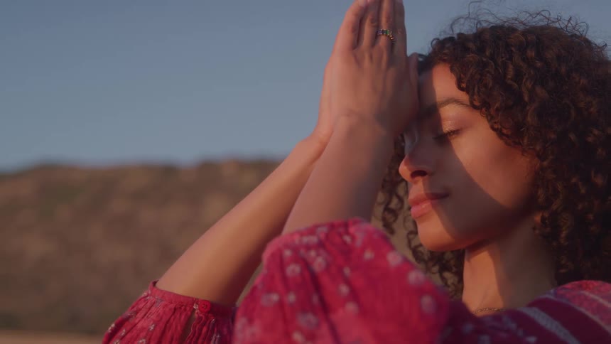 A woman with her eyes closed holding her hands together in a prayer pose with sunlight illuminating her face