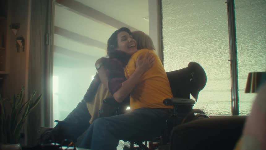 A young woman sits on a chair beside an elderly man in the living room watching a game they both suddenly become excited by the result and hug each other at midday