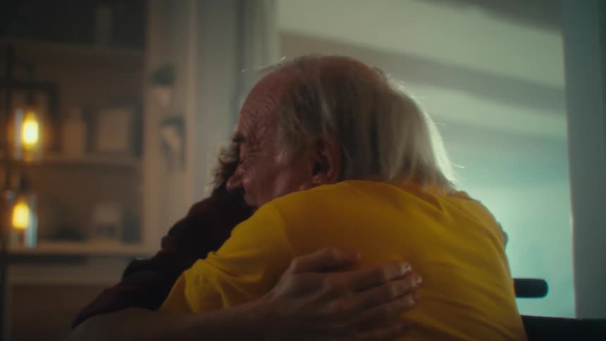 A man with white hair wearing a yellow shirt and a woman with brown hair wearing a plaid shirt are sitting together as they both look forward with excited expressions and the man raises his arms in celebration before embracing the woman