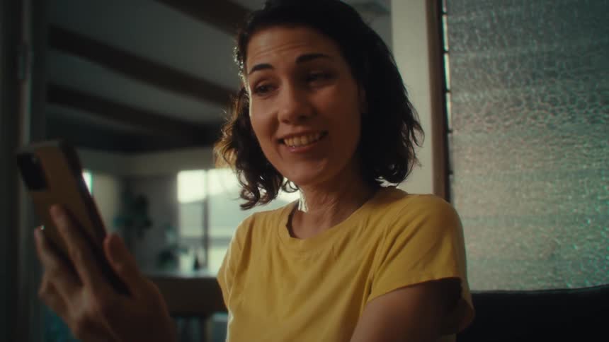 A woman with dark hair wearing a yellow shirt is sitting in a chair and looking down at a smartphone as she begins to smile and laugh while holding the phone in both hands