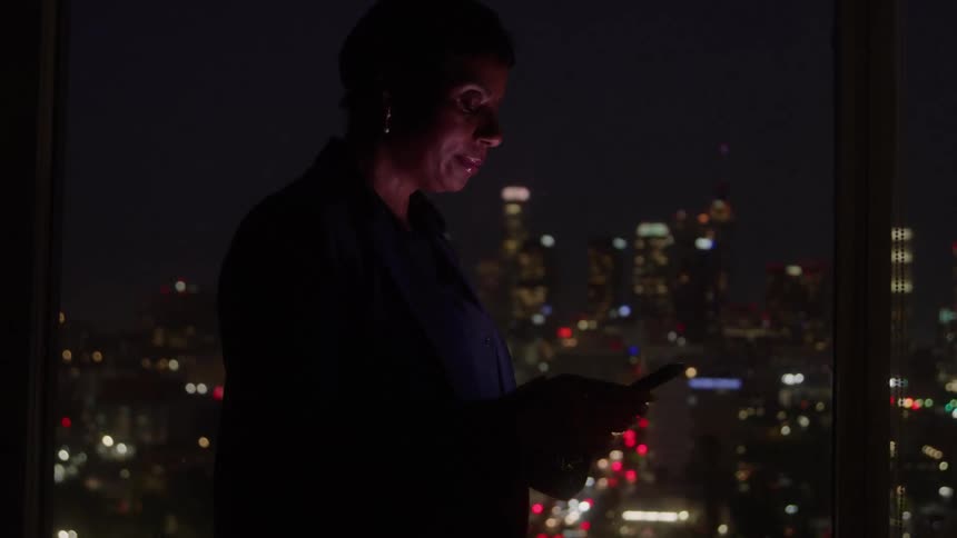 Woman using a cellphone while standing by a window overlooking the city at night