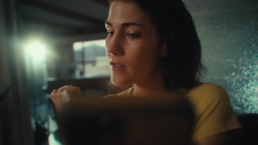 A woman with dark hair wearing a yellow shirt is looking down at a smartphone she holds in both hands as her facial expression changes from neutral to a big celebratory smile