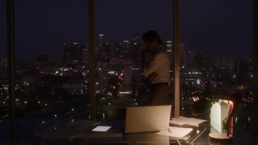 A man walks in front of a window at night looking at a city skyline with many bright lights near a closed laptop and some papers over a desk