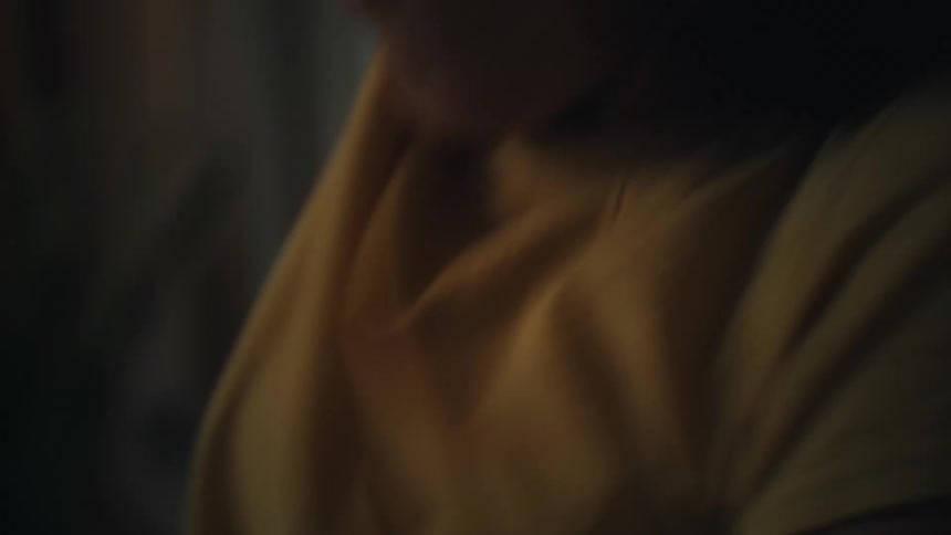 A woman with dark hair wearing a yellow shirt is sitting in a chair and playing a video game as she reacts with an excited and laughing expression