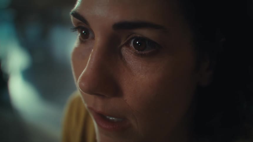 A close view shows a woman with dark hair and a light yellow shirt looking off to her right with a serious expression as her eyes shift and a bright light shines behind her