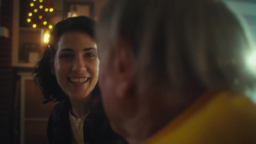 The video shows two people a man and a woman smiling and talking they are sitting indoors and the scene is slightly dark with some blurry warm lights in the background