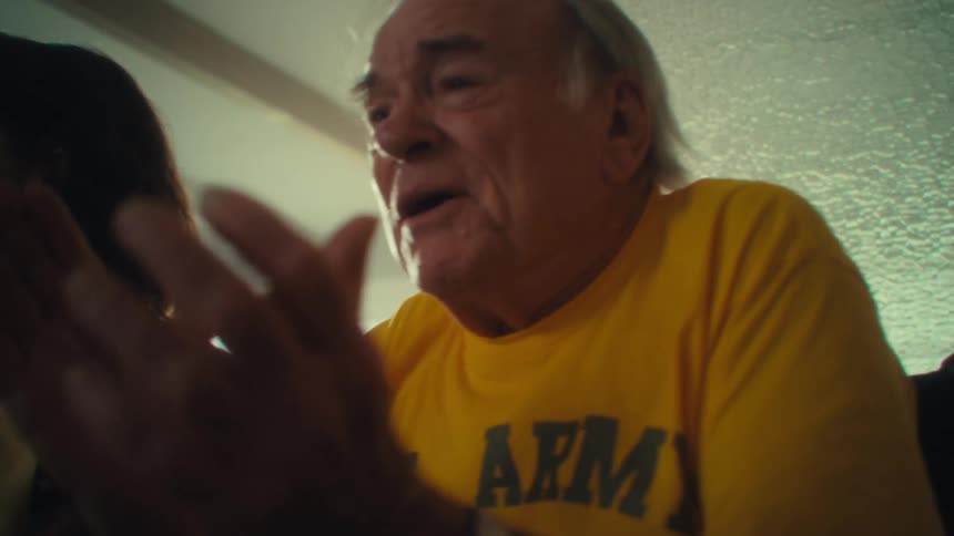 A woman with brown hair and a man with white hair and wearing a yellow shirt are sitting at a table together clapping their hands while smiling