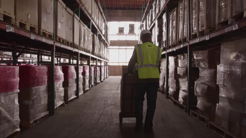 Worker wearing a reflective vest and hard hat pushing a hand truck with boxes in a warehouse