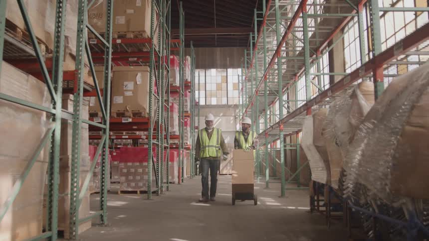 Two men inside a warehouse walk with one pushing a trolley loaded with shipping boxes both wearing hard hats and green vests as the warehouse is filled with shipping boxes at midday