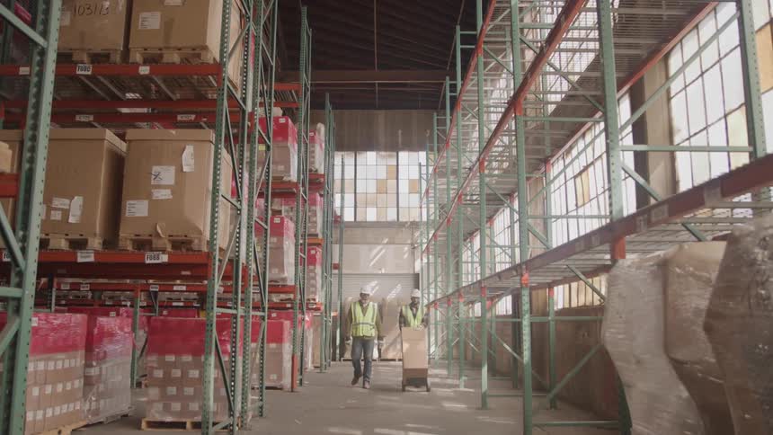 Two men wearing hard hats and green vests push a trolley down a warehouse aisle filled with shipping boxes at midday