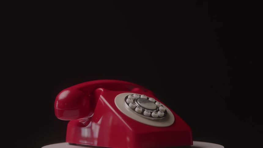 A medium view shows a red rotary phone with a white dial on a spinning white platform against a black background with the camera slowly moving backward