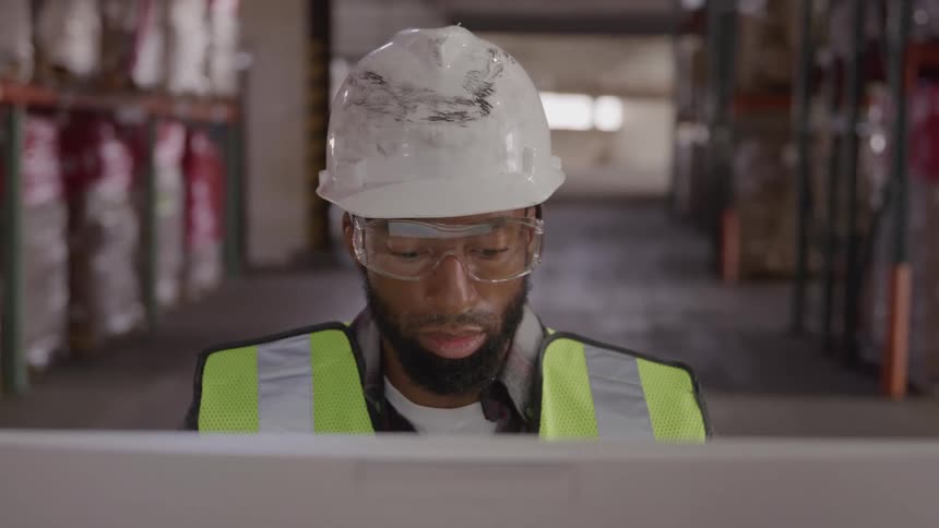 Man in helmet facing a computer in a warehouse