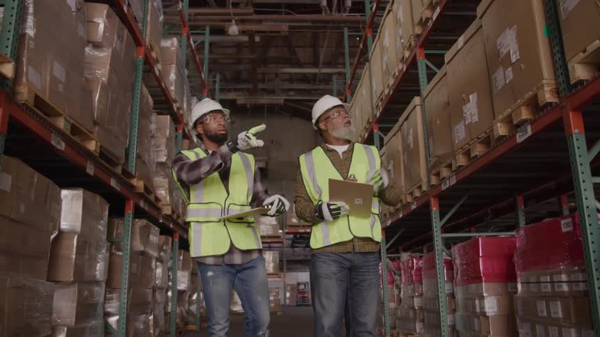 Two warehouse workers wearing safety vests and hard hats are walking between tall shelves of boxes they are holding clipboards with one worker pointing up and appearing to discuss the inventory with the other