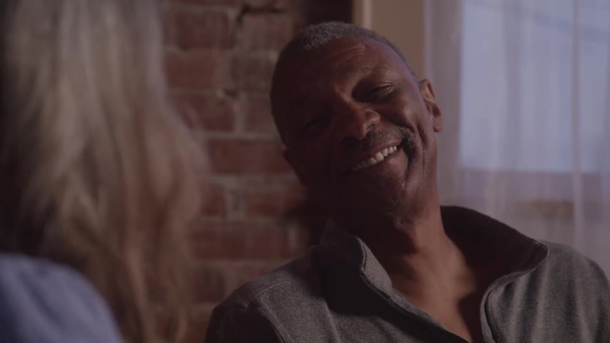A man with dark skin wearing a gray shirt is sitting next to a woman with gray hair looking at her and smiling with a brick wall and a window visible in the background