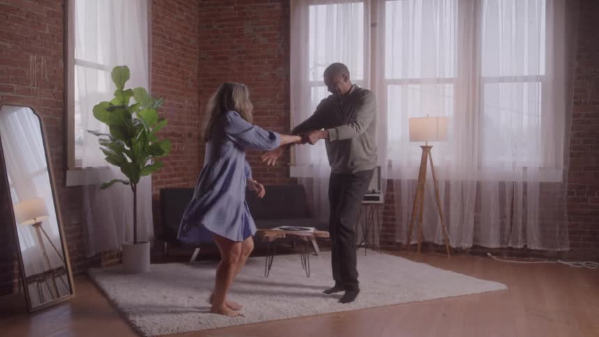 A man dances with his wife on a rug in their living room with a potted plant and a mirror beside the wall at midday