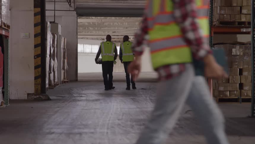 Two workers wearing reflective vests walking side by side inside a warehouse