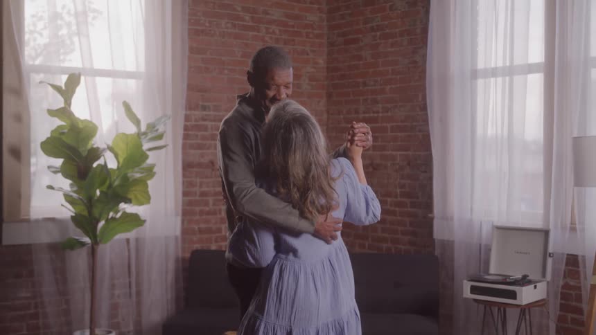 A couple dances to the rhythm of music slowly hugging each other indoors with a potted plant beside them at midday