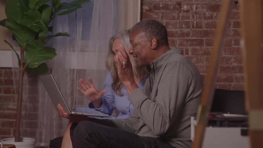 A older man and a woman are sitting together on a couch in a room with a brick wall both looking at a laptop and waving as if on a video call raising their hands with a big smile on their face