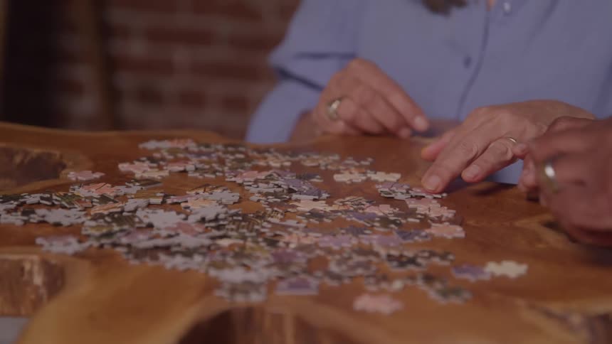 An older woman in a blue shirt works on assembling a jigsaw puzzle at a wooden table