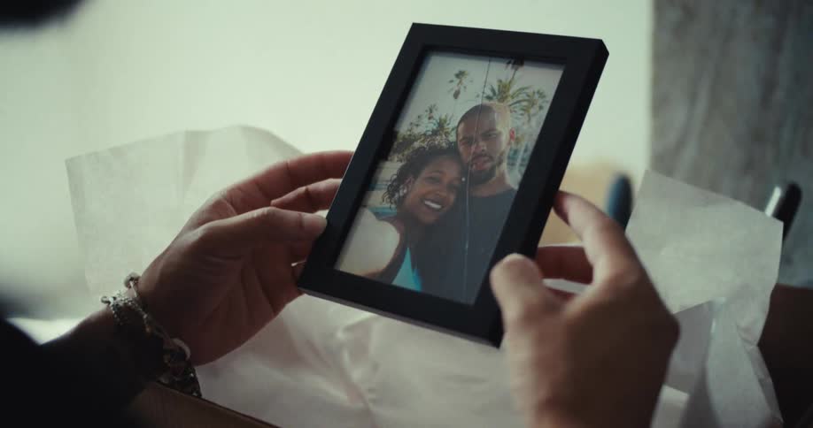 A person is unpacking a cardboard box labeled bed room and pulls out a black picture frame with a photograph of two people smiling in it one with their arm around the other