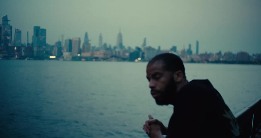 A bearded man stands in the foreground by the water at dusk with the new york city skyline and empire state building in the background