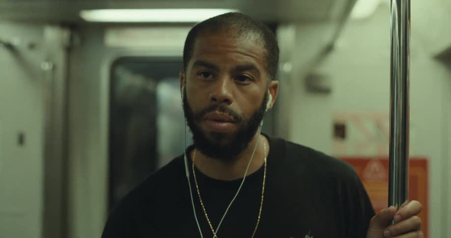 A man wearing earbuds and gold chains stands thoughtfully in a moving subway car holding onto a pole while commuting
