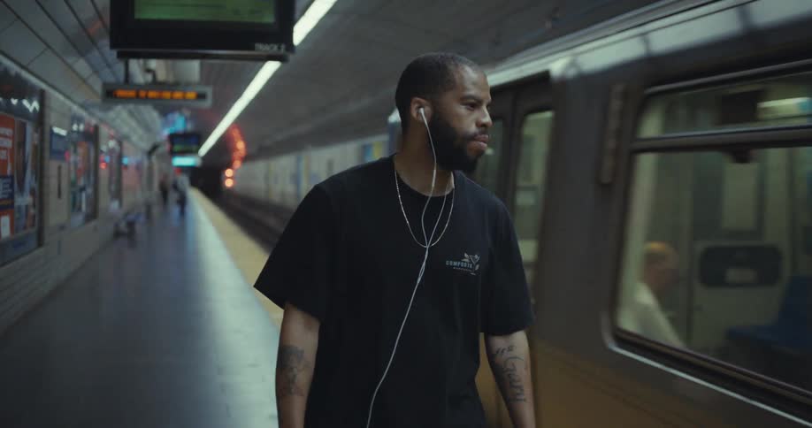 A bearded man with tattoos and wired earphones listens to music while walking on a dimly lit subway platform turning his head to watch the train as it rolls into the station