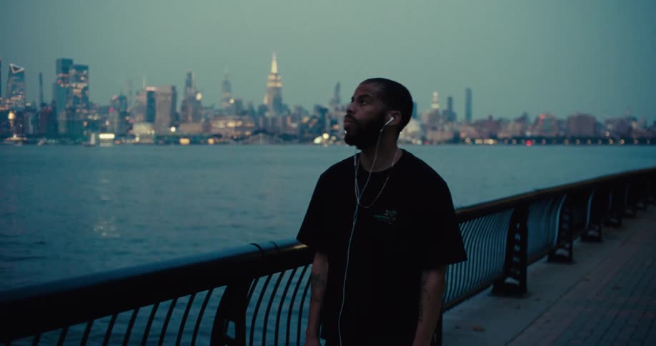 Man leans on the railing of a waterfront promenade with the city in the background