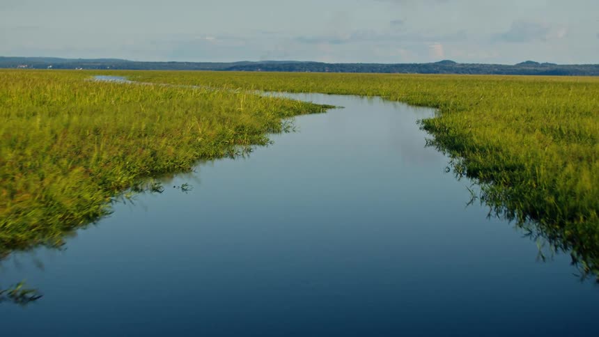 A low angle view of a wide dark body of water slowly tilts upward to reveal a clear river flowing through a field of green grass with a tree line and mountains in the distance