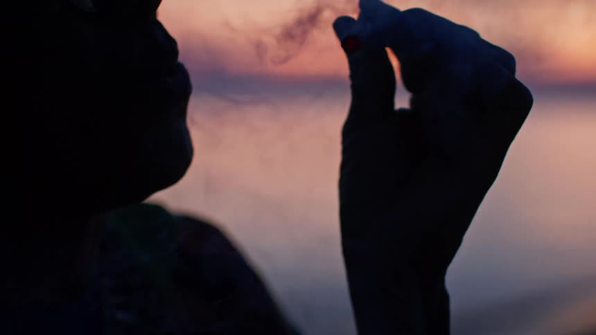 A closeup of a persons silhouette shows them lighting a cigarette with a lighter the flame briefly illuminates their hand and the side of their face before they out a puff of smoke