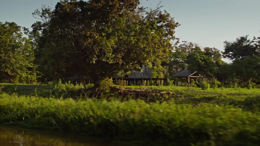 A long view of a calm river shows the reflection of a small wooden house sitting on a grassy riverbank as the camera slowly moves to the right revealing more of the rustic house and the trees surrounding it