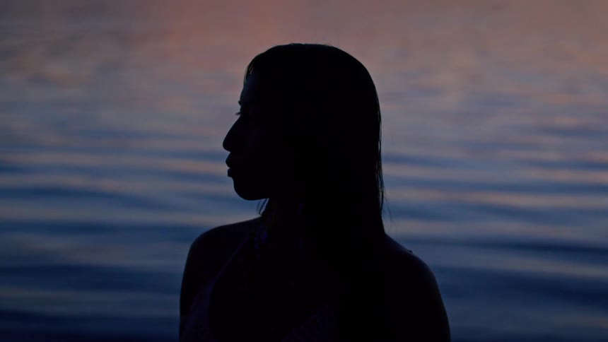 A womans silhouette is outlined against rippling water under the warm hues of sunset