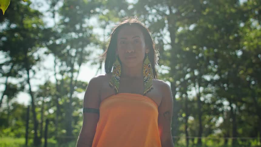 A woman staring straight ahead and standing outdoors and wearing an orange dress