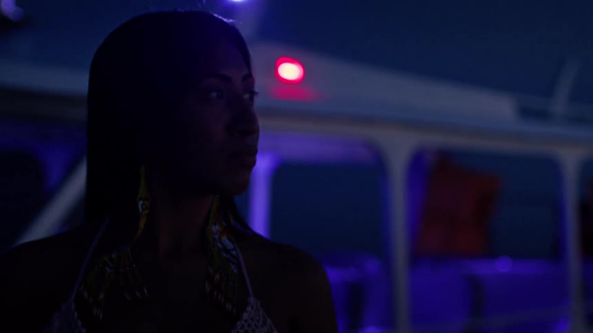 A woman in a white crocheted top stands in dim blue and purple lighting her face partly shadowed