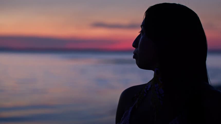 A womans silhouette faces the water at sunset with the horizon glowing in warm colors