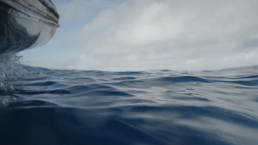 A low angle view from inside a small boat shows the waves of the dark blue ocean with a cloudy sky above as the camera dips into the water before resurfacing